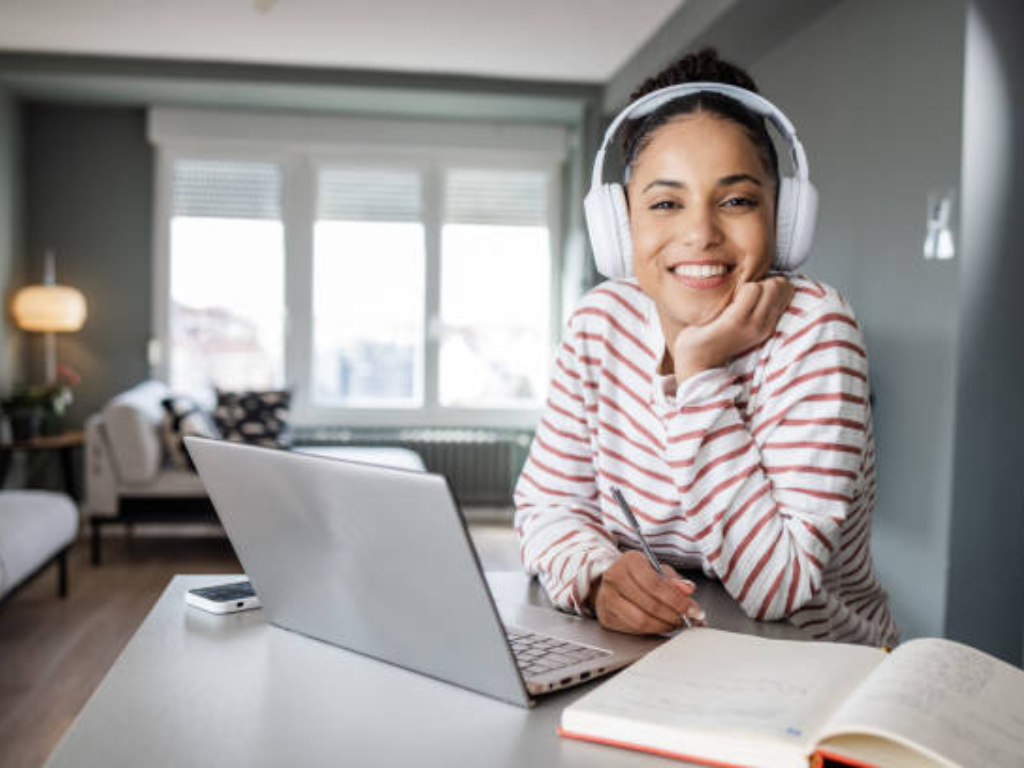 Mujer joven sonriendo mientras estudia con una computadora portátil y auriculares, en un entorno hogareño.